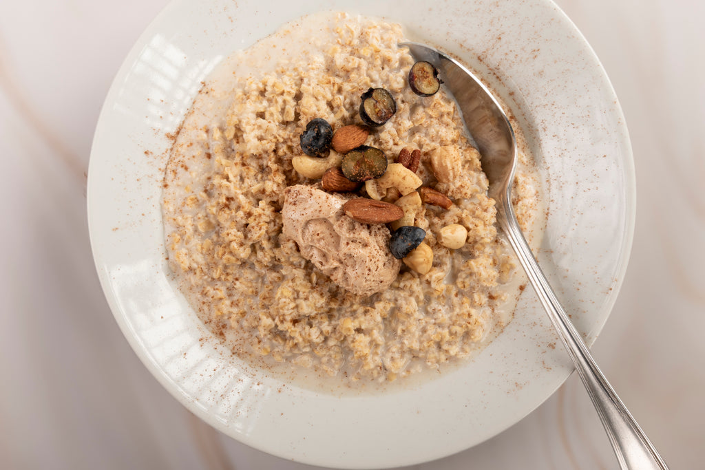 “Overhead view of a bowl of creamy porridge topped with a dollop of The Yorkshire Kitchen's Honey and Cinnamon butter, mixed nuts, and blueberries, lightly dusted with cinnamon. A metal spoon rests in the bowl on a light-coloured surface.