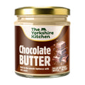 Glass jar of The Yorkshire Kitchen Chocolate Butter with a gold metal lid and rich brown label. The label features the brand name at the top and large white text reading ‘Chocolate Butter,’ alongside an illustration of stacked chocolate pieces. Additional text reads ‘Melt-in-the-mouth lightness with rich cocoa’ and ‘Dollop on waffles’ and ‘Spread on toast.’