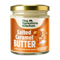 Glass jar of The Yorkshire Kitchen Salted Caramel Butter with a gold metal lid and orange label. The label features the brand name at the top and large white text reading ‘Salted Caramel Butter,’ alongside an illustration of stacked caramel cubes sprinkled with salt. Additional text reads ‘Smooth, sweet and utterly moreish’ and ‘Dollop on pancakes’ and ‘Stir into hot chocolate.’