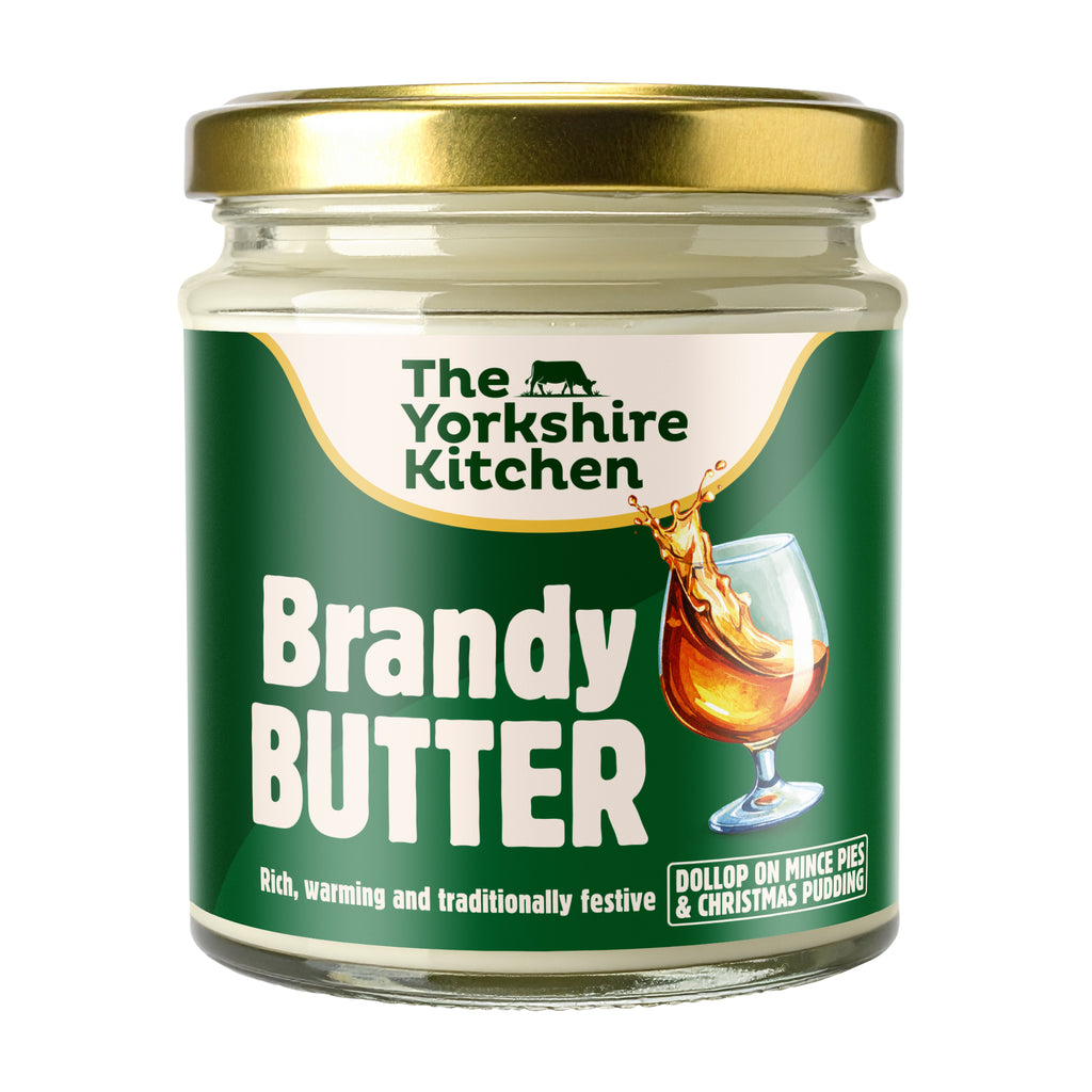 Glass jar of The Yorkshire Kitchen Brandy Butter with a gold metal lid and green label. The label features the brand name at the top and large white text reading ‘Brandy Butter,’ alongside an image of a glass of brandy splashing. Additional text reads ‘Rich, warming and traditionally festive’ and ‘Dollop on mince pies & Christmas pudding.