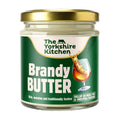 Glass jar of The Yorkshire Kitchen Brandy Butter with a gold metal lid and green label. The label features the brand name at the top and large white text reading ‘Brandy Butter,’ alongside an image of a glass of brandy splashing. Additional text reads ‘Rich, warming and traditionally festive’ and ‘Dollop on mince pies & Christmas pudding.