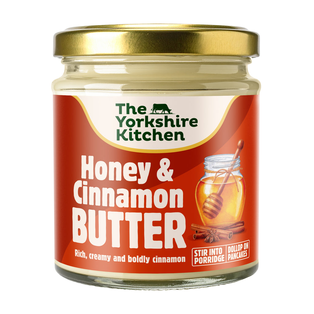 Glass jar of The Yorkshire Kitchen Honey & Cinnamon Butter with a gold metal lid and warm red label. The label features the brand name at the top and large white text reading ‘Honey & Cinnamon Butter,’ alongside an illustration of a jar of honey with a dipper and cinnamon sticks. Additional text reads ‘Rich, creamy and boldly cinnamon’ and ‘Stir into porridge’ and ‘Dollop on pancakes.’