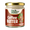 Glass jar of The Yorkshire Kitchen Coffee Butter with a gold metal lid and deep brown label. The label features the brand name at the top and large white text reading ‘Coffee Butter,’ alongside an illustration of coffee beans with green leaves. Additional text reads ‘Gentle coffee tones with a warm finish’ and ‘Spread on a croissant’ and ‘Smear over a crumpet.'