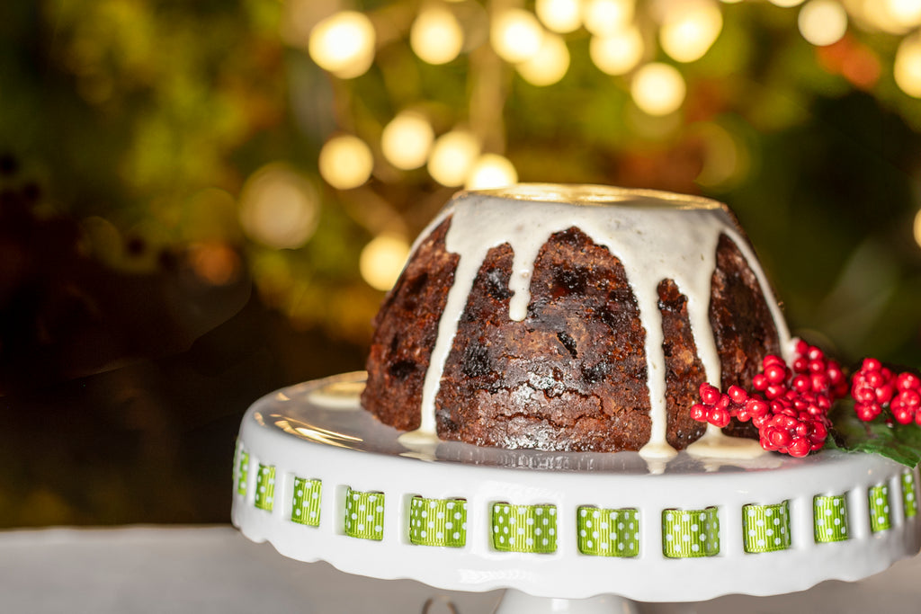 Traditional Christmas pudding topped with white icing dripping down the sides, displayed on a white cake stand decorated with a green ribbon detail. The pudding is garnished with bright red berries, with warm golden lights softly blurred in the background.
