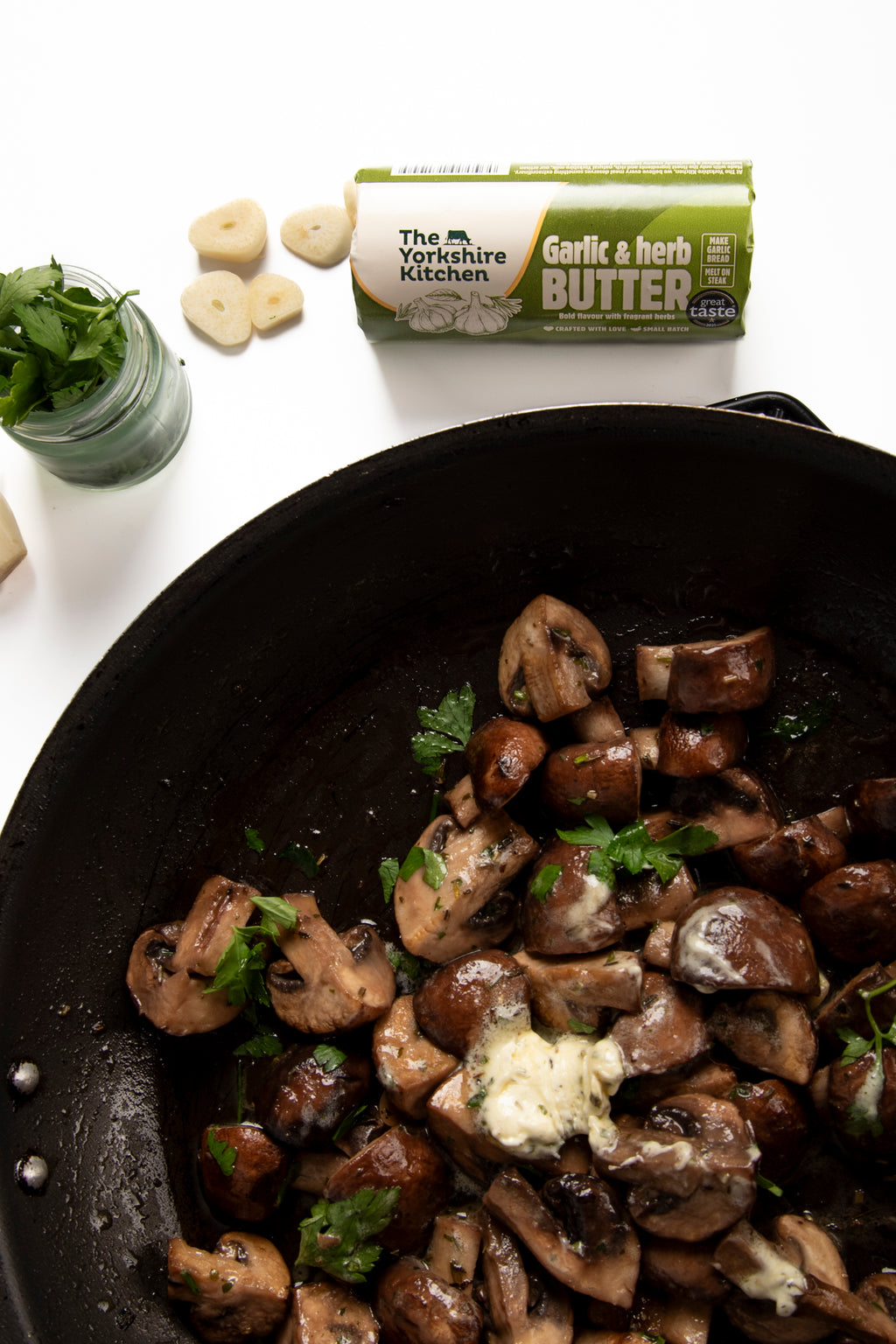 Overhead food scene showing sautéed mushrooms in a black pan topped with a melting dollop of garlic and herb butter and sprinkled with chopped parsley. In the background on a white surface are a wrapped roll of The Yorkshire Kitchen Garlic & Herb Butter, sliced garlic cloves, and a small jar of fresh herbs.
