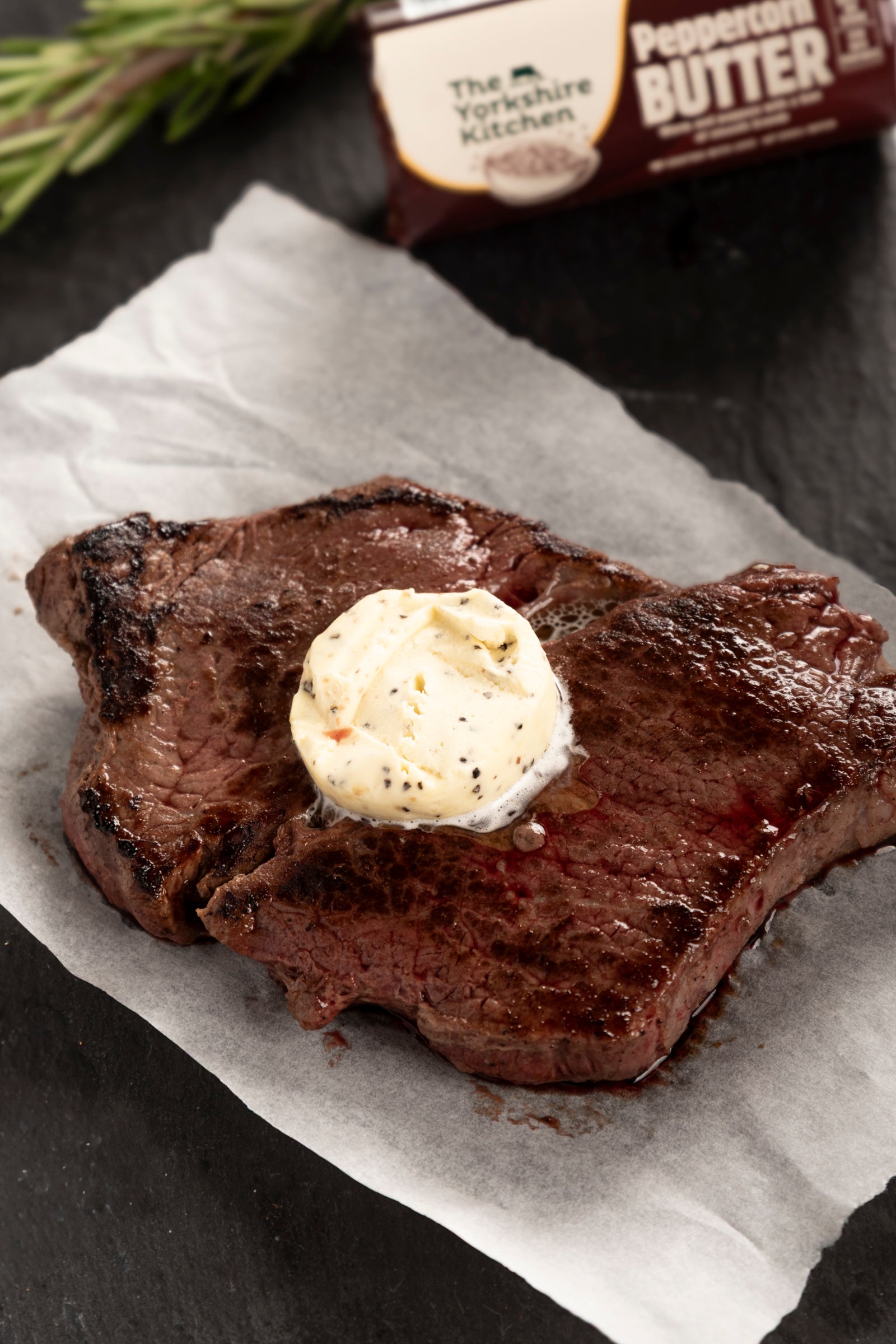 Grilled steak resting on parchment paper with a melting round of peppercorn butter on top. In the background, a wrapped roll of The Yorkshire Kitchen Peppercorn Butter is partially visible on a dark surface with a sprig of fresh rosemary.